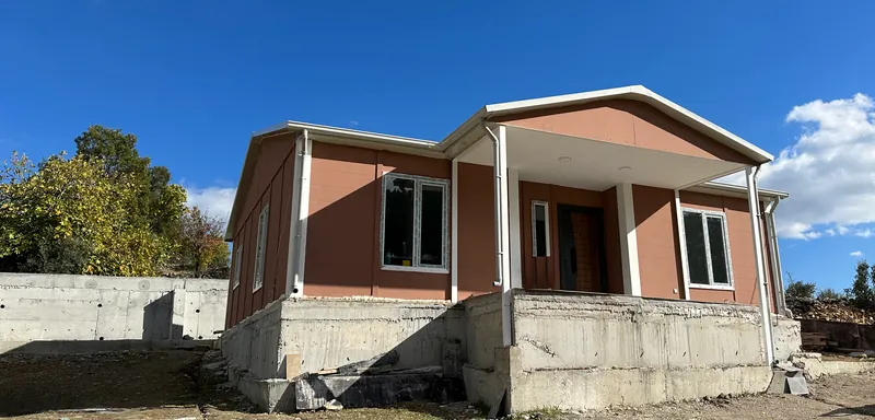 Terrakota panel duvarlı, beyaz profilli ve yükseltilmiş beton temel üzerinde kapalı verandalı yeni evin önden görünümü