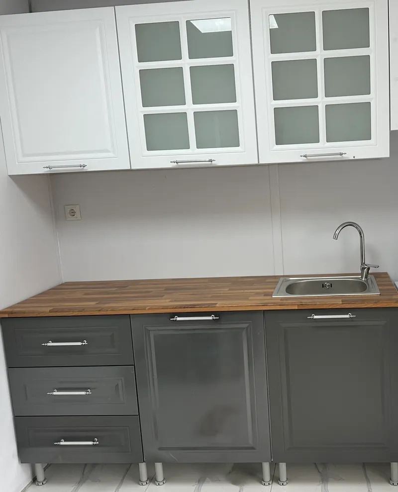 Beyaz camlı üst dolaplar, gri alt modüller ve evyeli ahşap tezgâhtan oluşan küçük mutfak